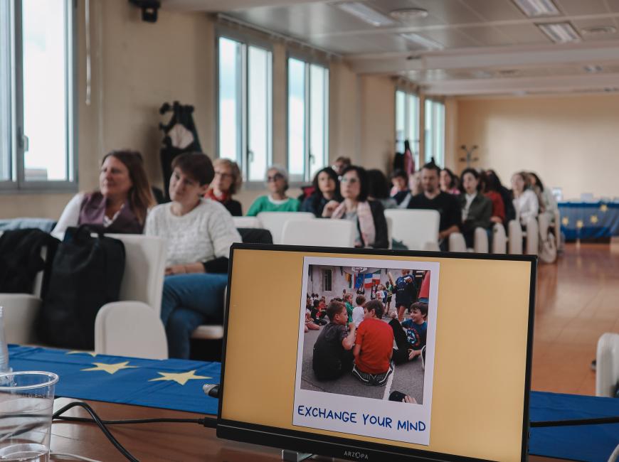 Fotografia che inquadra il computer e dietro il pubblico presente all'evento Europa a Scuola.