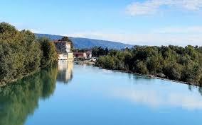 in primo piano un fiume con vegetazione sulle sponde. in fondo edifici storici